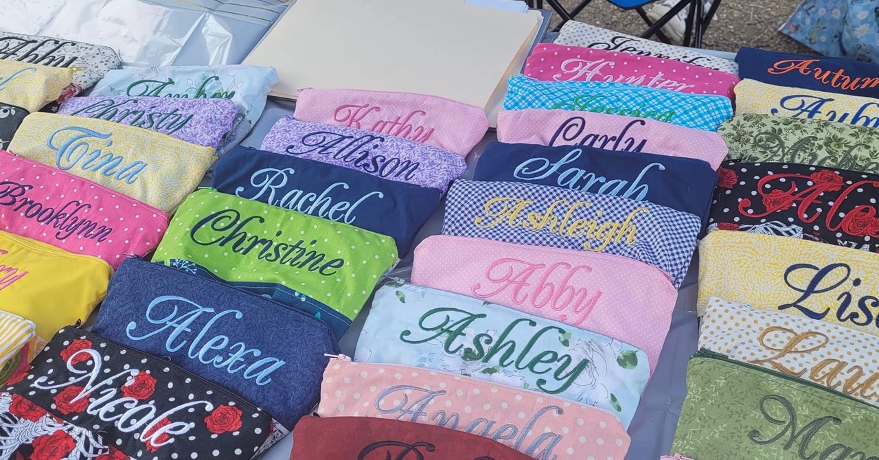 A craft-fair table of personalized embroidered zip pouches, each stitched with a different name